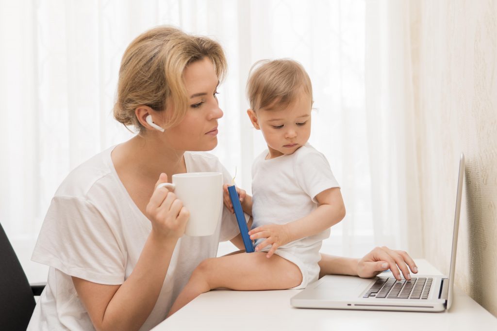 Eine entspannte Mama mit einem Laptop auf dem Tisch, Kaffee trinkend und Kind dabei